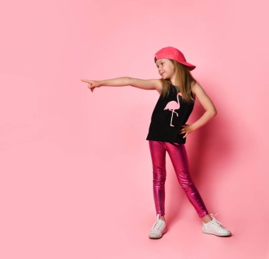 Kendine güvenen, beyzbol şapkası takmış, eli kalçasında poz veren küçük bir kız. Tam uzunlukta çekim pembe üzerinde izole edildi, kopyala boşluk