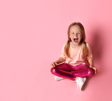 Tişörtlü, taytlı ve spor ayakkabılı küçük bir kadın. Yukarı bakıyor, pembe arka planda yerde oturuyor..