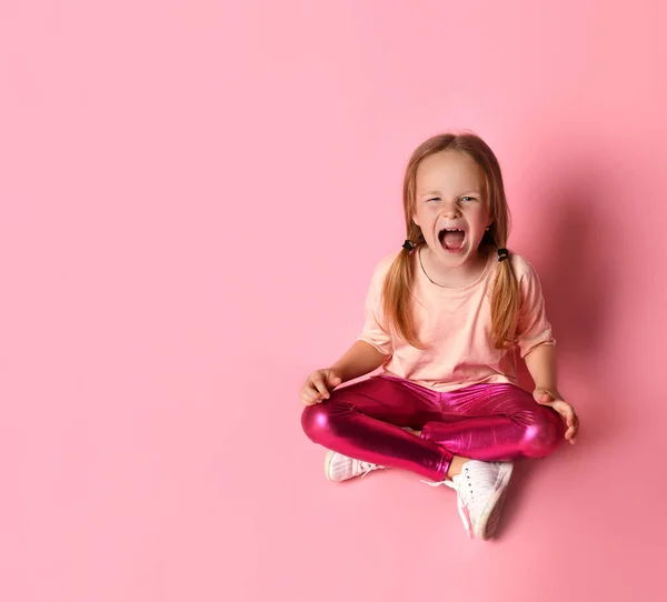 Pequeña hembra en camiseta, polainas y zapatillas. Ella mirando hacia arriba, sentada en el suelo sobre fondo rosa. — Foto de Stock