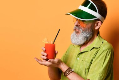 Yeşil güneşlik, gömlek ve güneş gözlüklü sakallı adam. Bir bardak sıkılmış meyve suyu, sıkılmış yumruk, turuncu arka planda poz.