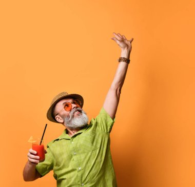 Sakallı, şapkalı, yeşil gömlekli, güneş gözlüklü bir erkek. Dans eder gibi elini kaldırdı. Elinde meyve suyu bardağıyla turuncu arka planda poz verdi.