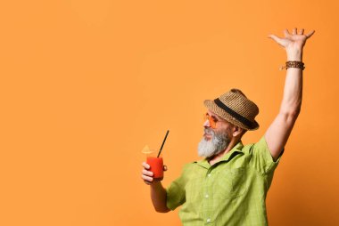 Sakallı, şapkalı, yeşil gömlekli, güneş gözlüklü bir erkek. Dans eder gibi elini kaldırdı. Elinde meyve suyu bardağıyla turuncu arka planda poz verdi.