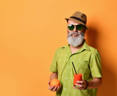 Şapkalı yakışıklı bir adam elinde bir portakal suyu ve bir bardak taze sıkılmış meyve suyu tutuyor. Siyah bir tüple turuncu stüdyo arka planında yan poz veriyor. Kopyalama alanını kapat