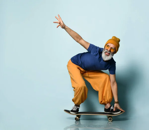 Bearded elderly man in t-shirt, sunglasses, orange pants, hat, gumshoes. Riding black skateboard, posing on blue background
