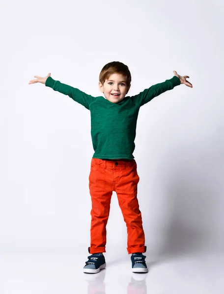 Malý brunet model v zeleném svetru, oranžové kalhoty, modré tenisky. Zvedl ruce a tvářil se rozradostněně, pózoval izolovaně na bílém — Stock fotografie
