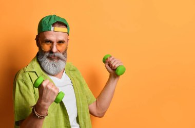 Büyükbaba yeşil şapkalı ve gömlekli, beyaz tişörtlü, bilezik takıyor. Gülümsüyor, halterlerle egzersiz yapıyor, turuncu arka planda poz veriyor.
