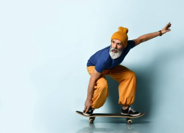 Tişörtlü, turuncu pantolonlu, şapkalı, spor ayakkabılı yaşlı bir adam. Siyah bir kaykaya biniyor, ona bakıyor, mavi arka planda poz veriyor.