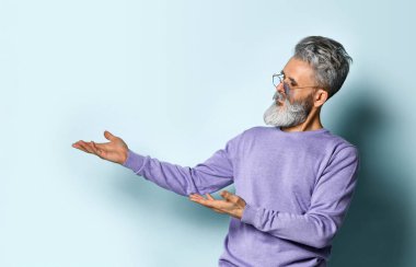 Gri saçlı, sakallı, güneş gözlüklü, mor kazaklı yaşlı bir adam. Mavi arka planda elle poz vermek. Kapat.