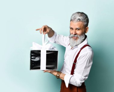 Beyaz gömlekli, kahverengi pantolonlu, askılı, bilezik takan yaşlı bir erkek. Elinde gümüş bir hediye kutusu var, mavi arka planda yan poz veriyor.