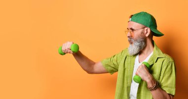 Yeşil şapkalı, tişörtlü, beyaz tişörtlü, güneş gözlüklü yaşlı bir erkek. İki dambılı boks yapıyor, turuncu arka planda yan poz veriyor.