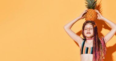 Afrikalı örgü saçlı, renkli mayo giyen genç bir kız. Gözleri kapalıydı, kafasında ananas vardı, sarı arka planda poz veriyordu.