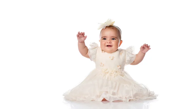 Vacker baby barn i en festlig klänning och ett pannband, sitter på golvet och glatt höjde händerna. skott på en vit bakgrund. — Stockfoto