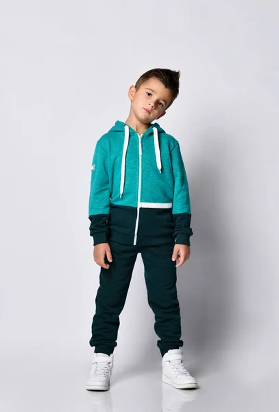 Liten brunett unge i färgglada träningsoverall och sneakers på en grå bakgrund. — Stockfoto