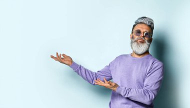 Gri saçlı, sakallı, güneş gözlüklü, mor kazaklı olgun bir adam. Mavi arka planda poz verirken elleriyle bir şey gösteriyor. Kapat.