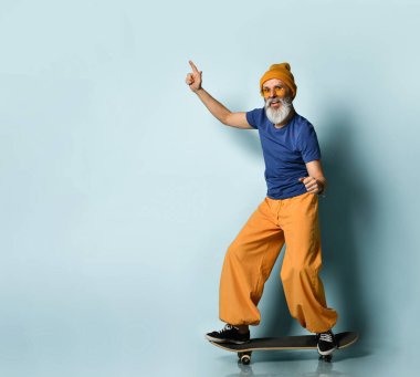 Tişörtlü, güneş gözlüklü, turuncu pantolonlu, şapkalı, spor ayakkabılı yaşlı bir adam. Siyah kaykay sürmek, yumruklarını göstermek, mavi arka planda poz vermek.