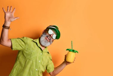 Büyükbaba yeşil güneşlik, gömlek, güneş gözlüğü takıyor. Kokteyl şişesini ananas şeklinde tutarak, turuncu arka planda poz vererek.