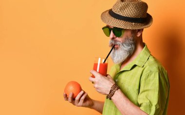 Şapkalı, yeşil gömlekli, güneş gözlüklü yaşlı bir adam. Turuncu arka planda durarak, portakal suyu tutarak ve tüple taze meyve suyu içerek.