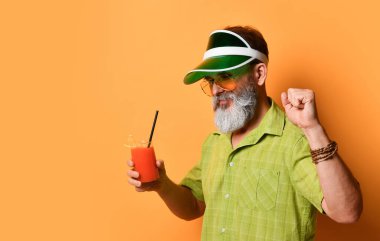 Yeşil güneşlik, gömlek ve güneş gözlüklü sakallı adam. Bir bardak sıkılmış meyve suyu, sıkılmış yumruk, turuncu arka planda poz.