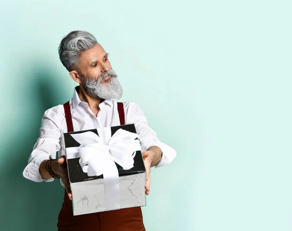 흰 셔츠에 갈색 바지에 멜빵 바지에 팔찌를 매고 있는 나이든 남자. 은으로 된 선물 상자를 들고 파란 배경에 옆으로 누워 있는 모습 — 스톡 사진