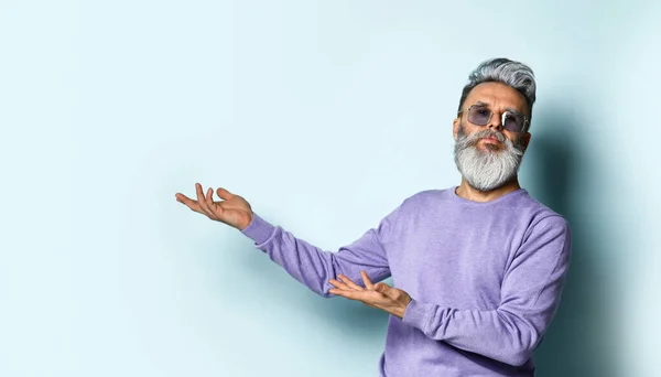 Homme mûr barbu aux cheveux gris, lunettes de soleil, pull violet. Montrant quelque chose par ses mains, posant sur fond bleu. Gros plan — Photo