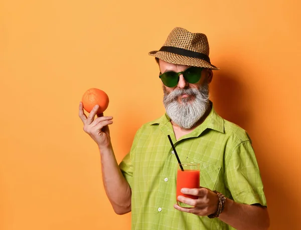 Aged man in hat, green shirt, sunglasses. Smiling, holding an orange, glass of fresh juice with tube, posing on orange background