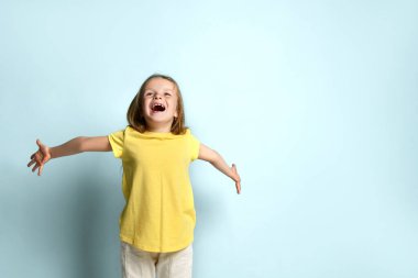 Sarı tişörtlü ve beyaz pantolonlu küçük bir kadın. Ellerini ayırıp çığlık atıyor, turkuaz arka planda poz veriyor. Kapat.