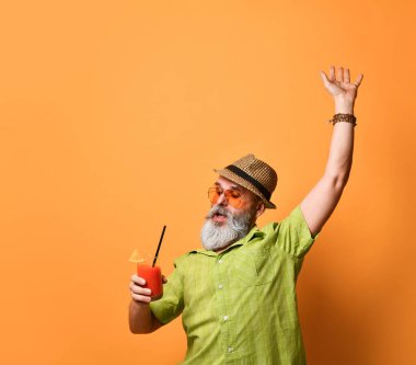 Sakallı, şapkalı, yeşil gömlekli, güneş gözlüklü bir erkek. Dans eder gibi elini kaldırdı. Elinde meyve suyu bardağıyla turuncu arka planda poz verdi.