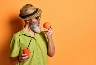 Şapkalı, yeşil gömlekli, güneş gözlüklü yaşlı bir erkek. Elinde bir bardak sıkılmış meyve suyu ile portakal arkaplanda poz vererek portakalı ısırıyor.