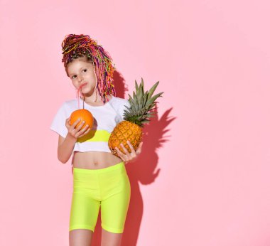 Kafasında çok renkli at kuyrukları olan sevimli küçük bir kız elinde ananas ve portakalla mutlu bir şekilde gülümsüyor. Kesilmiş portre pembeye izole edilmiş, kopyalama alanı. Çocukluk, duygular, yaz.