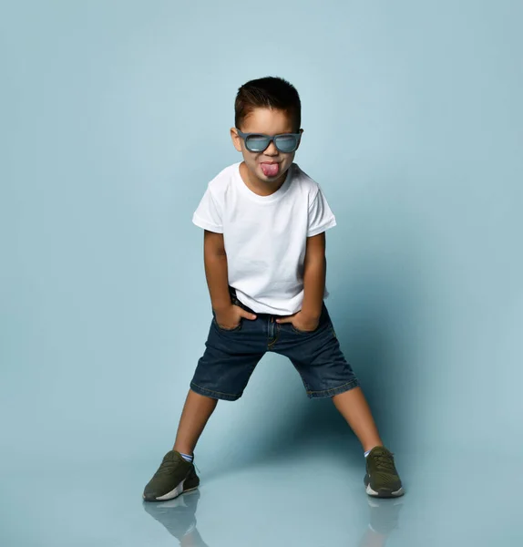 Маленький хлопчик в сонцезахисних окулярах, біла футболка, джинсові шорти та кросівки хакі. Усміхаючись, кладіть руки в кишені, позує на синьому фоні — стокове фото