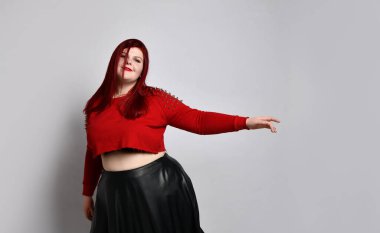 Kırmızı kazıklı, siyah sutyen ve deri etekli obez kadın. Dans ediyor, beyaz arka planda yalnız poz veriyor.