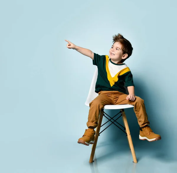 Dítě v barevném tričku, hnědé kalhoty. Usmívat se, na něco ukazovat, sedět na bílém křesle na modrém pozadí — Stock fotografie