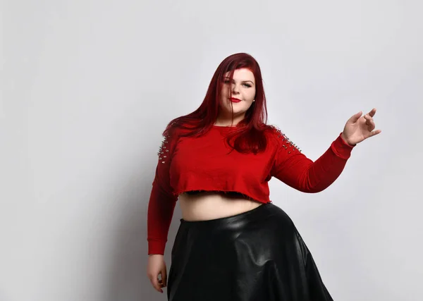 Femme gingembre obèse en haut à pointes rouges, soutien-gorge noir et jupe en cuir. Elle danse, posant isolée sur fond de photo blanche — Photo