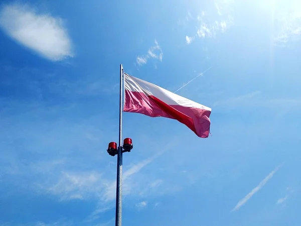 Le drapeau blanc-rouge polonais se développe contre le ciel bleu et les nuages blancs par une journée ensoleillée. traces de l'avion dans le ciel . — Photo