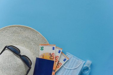 Üzerinde kırmızı kurdele, maske, dezenfektan, pasaport, para-avro ve mavi sandalet olan üst görüş şapkası. Coronavirüs 'te tatil. Yaz düzlüğü, kopyalama alanı, seyahat ve boş zaman kavramı olan kompozisyon.