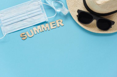 Hasır şapkalı, güneş gözlüklü, yaz tahtadan harflerle dezenfekte edilmiş tıbbi maske. Seyahat ve eğlence anlayışı. Denize uçuşlar, koronavirüs sırasında dinlenme. Tatil Mavinin üstünde