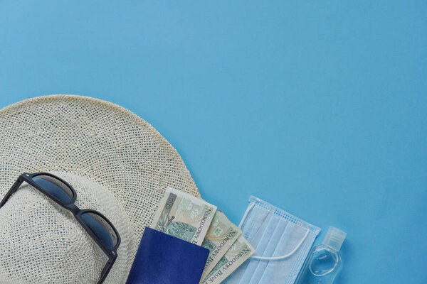 Top view hat with red ribbon, mask, sanitizer, passport, money-zloty and red flip flop sandals on blue.Vacation on coronavirus. Summer flatley, composition with copy space, travel and leisure concept.