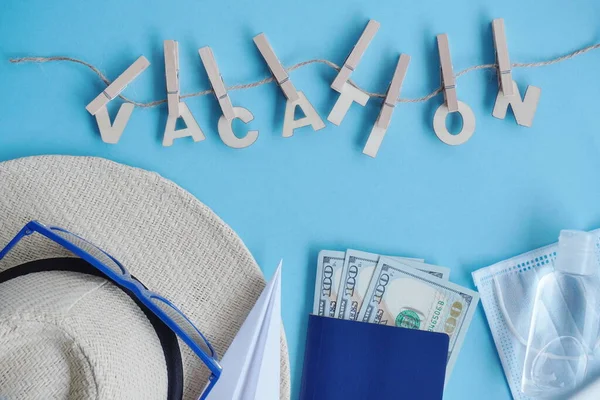 Top view hat,mask, sanitizer, passport, money, paper airplane -dollars on blue. Relax on coronavirus. Summer flatley, composition with copy space,travel and leisure concept. Wooden word Vacation