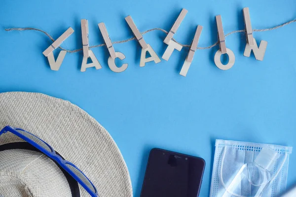 Top view hat,mask, sanitizer, mobile phone, paper airplane on blue. Relax on coronavirus. Summer flatly, composition, copy space, safe travel and leisure concept.Wooden word Vacation