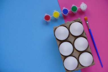 Mavi ve pembe arka planda altı yumurta tepsisi ya da kutusu, çok renkli boya ve fırçalar. Paskalya 'da çocukların yaratıcılığına göre ayarlandı. Asgari paskalya konsepti. Sembol Mutlu Paskalya Düzlüğü