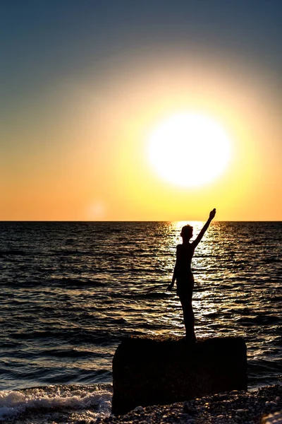 海の上の夕日の背景に人間の姿のシルエット 男は大きな岩の上に立っていて その上で海の波は壊れ 沈みゆく太陽に波が来る — ストック写真