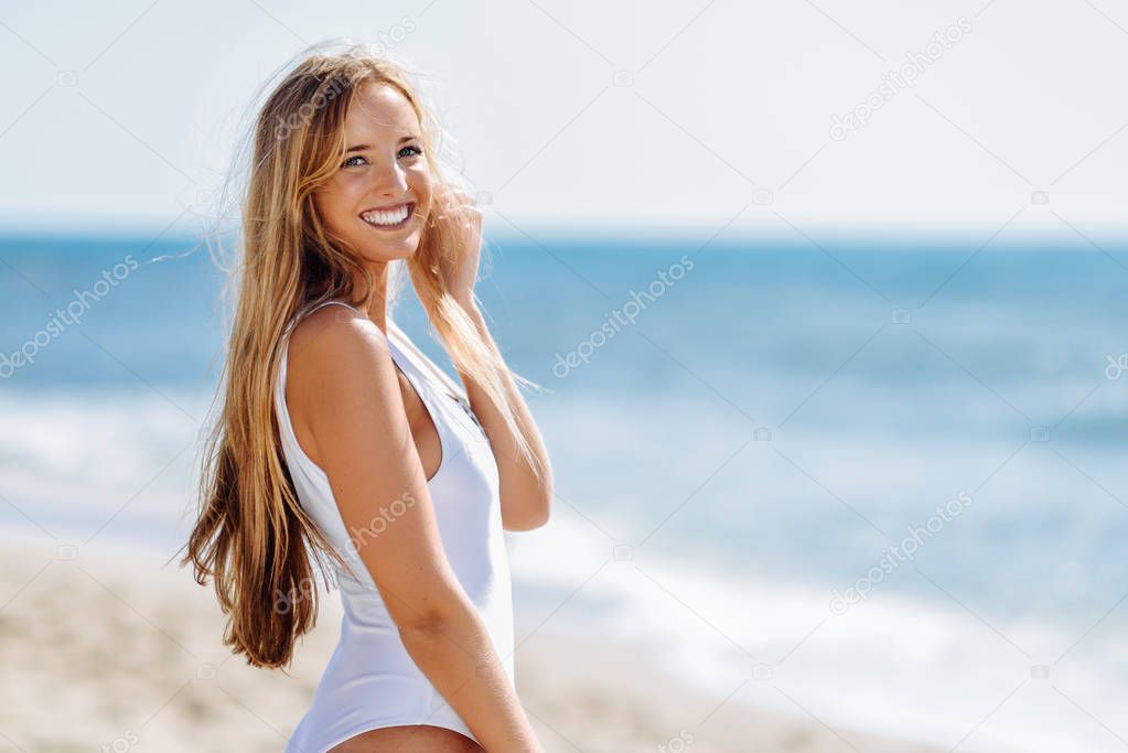 Young blonde woman with beautiful body in white swimsuit on a tropical beach. Caucasian female with straight long hairstyle smiling.