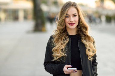 Blonde woman texting with her smart phone in urban background. Beautiful young girl wearing black jacket walking in the street. Pretty russian female with long wavy hair hairstyle.