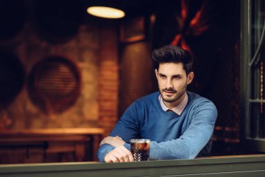 Modern bir barda kayıp bakışla mavi kazaklı genç adam. Bir soda içme bir pencere yakınında modern saç modeli ile düşünceli adam.