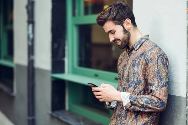 年轻的微笑男子穿着休闲服装看着他的智能手机在街上 胡子和现代发型在城市背景的家伙 — 图库照片