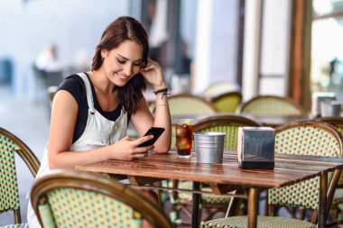 Akıllı telefon gülümseyen kullanarak kentsel kafe oturan mavi gözlü kız. Beyaz kot elbise kahverengi dalgalı saç modeli ile mutlu bir kadın. Yaşam tarzı kavramı.