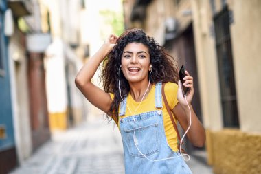 Çekici Kuzey Afrika kadın açık havada kulaklık ile müzik dinlemek. Kentsel arka planda kıvırcık saç modeli ile rahat kıyafetler içinde Arap kız.