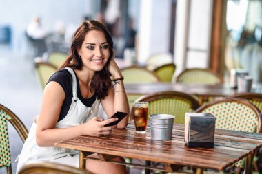 Akıllı telefon gülümseyen kullanarak kentsel kafe oturan mavi gözlü kız. Beyaz kot elbise kahverengi dalgalı saç modeli ile mutlu bir kadın. Yaşam tarzı kavramı.