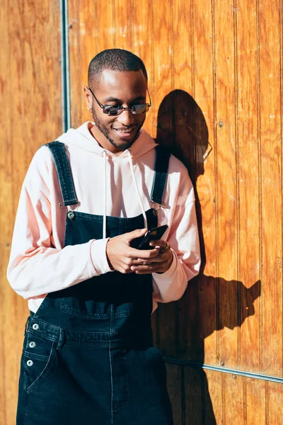 Young black man wearing casual clothes and sunglasses using smart phone against a wooden background. Millennial african guy with bib pants outdoors smiling