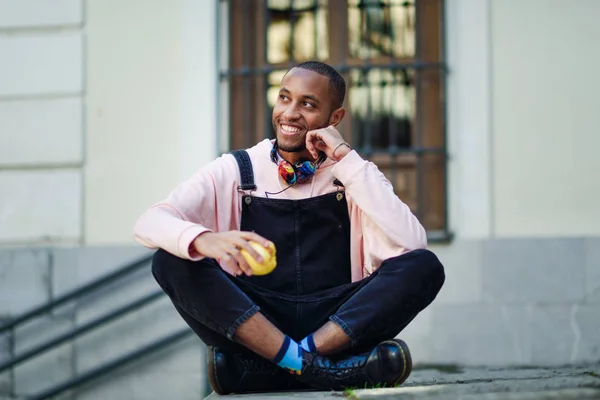 Young black man eating an apple sitting on urban steps. Lifestyle concept.